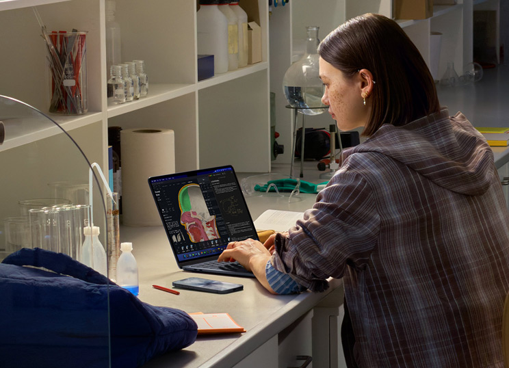 Eine Person sitzt in einem Labor und benutzt ein MacBook, auf dem mehrere Apps angezeigt werden.