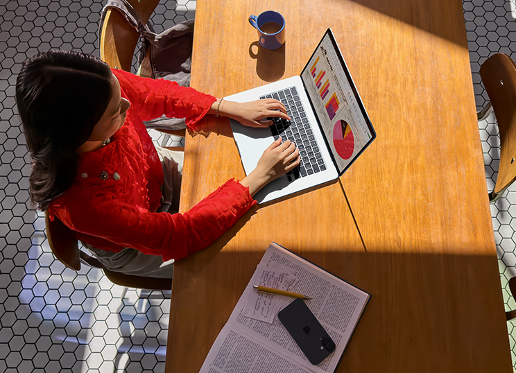 A person using MacBook at a table, with a mug, smartphone and papers nearby.