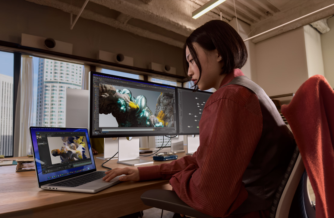 デスクで作業する人物。MacBook Proが外部ディスプレイに接続されており、両方の画面に3Dレンダリングが表示されている。窓の外には建物が見える