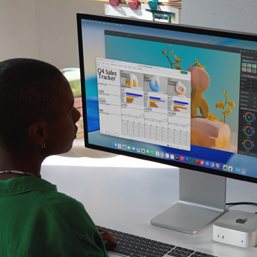 Una persona sentada trabaja en una hoja de cálculo de Excel titulada "Registro de ventas del cuarto trimestre" en un iMac y un Mac mini. Detrás de la hoja de cálculo de Excel hay una ventana de Pixelmator Pro y el fondo de macOS Tahoe.