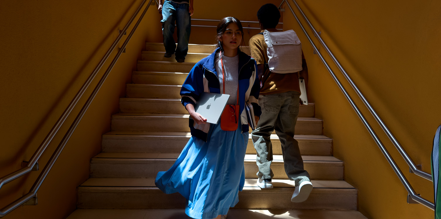 Una estudiante baja las escaleras. Lleva una MacBook y unos libros en las manos.