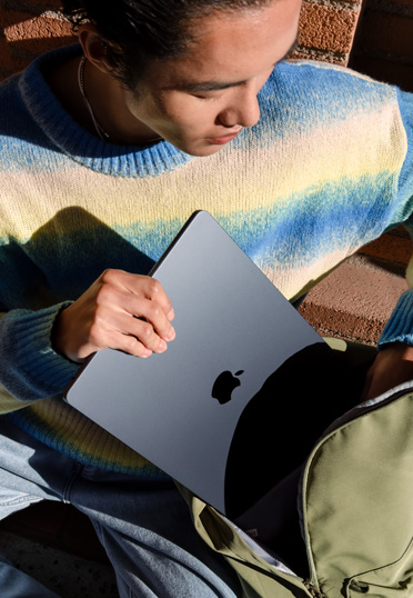 Un estudiante sentado al aire libre saca una MacBook cerrada de su mochila.