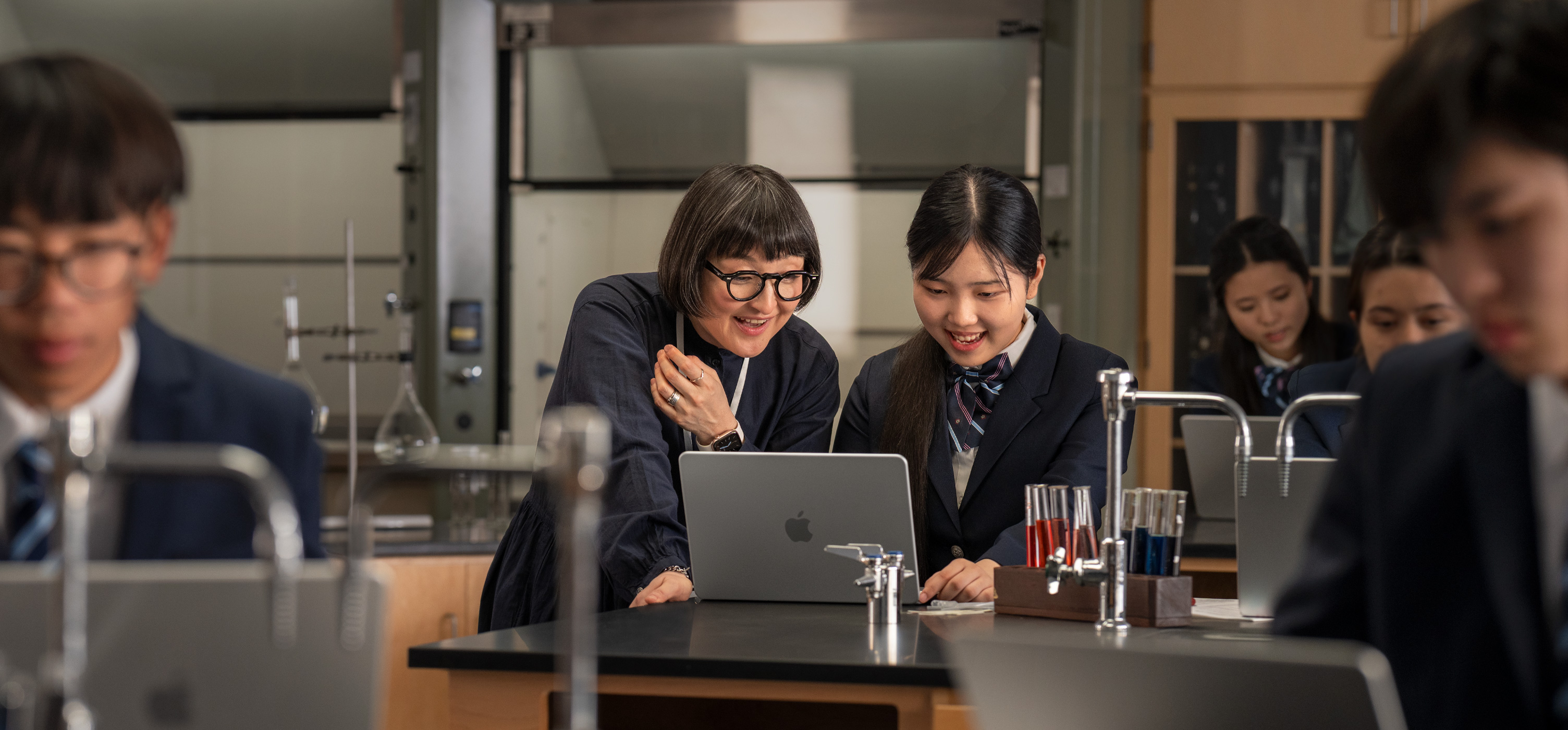 一位學生和一位教師在實驗室環境下互動，桌上放著打開的 MacBook，周圍滿佈試管和設備