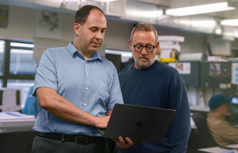Two men at a workstation look at a MacBook together, one holding it while the other points at the screen.