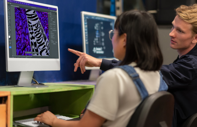 Two students sit in front of iMac monitors displaying colorful models, one pointing at the screen.