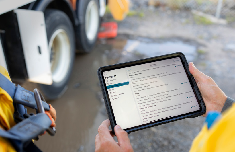 Hands hold an iPad with the HazChat app open which displays Job Details, Planning, Hazards and Controls, while a person stands at a worksite by a mud puddle