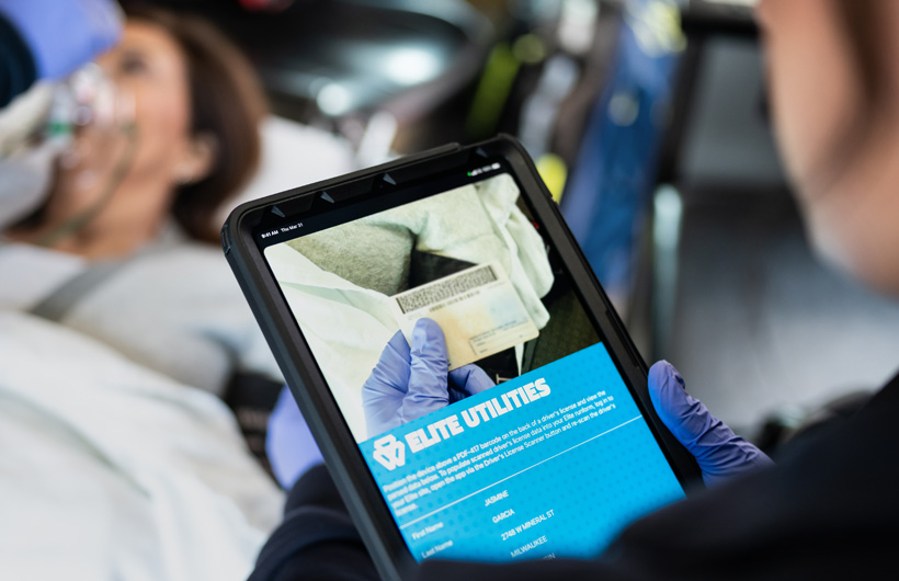 O app Elite Utilities está aberto no iPad, e um paramédico escaneia a carteira de motorista de uma paciente, que está deitada na maca