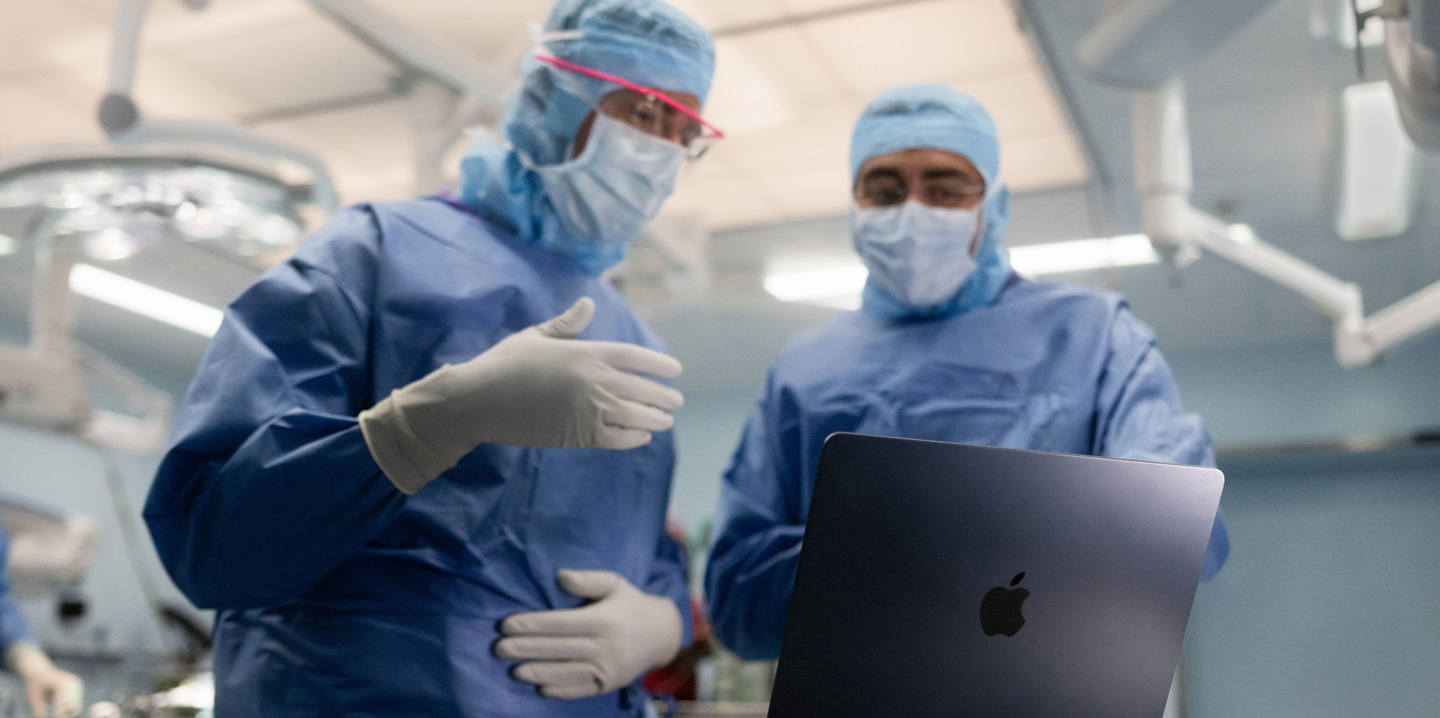Dois cirurgiões olham para um MacBook na sala de cirurgia, e um gesticula com a mão enquanto o outro observa