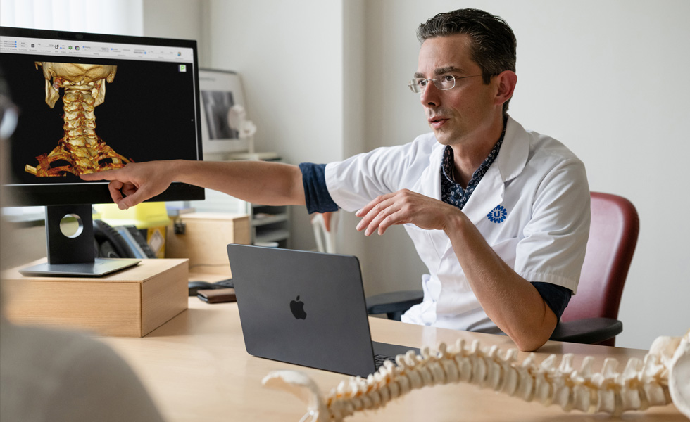 Profissional de saúde a apontar para uma representação em 3D dos ossos da parte de trás do pescoço no Studio Display, com o MacBook pousado na secretária
