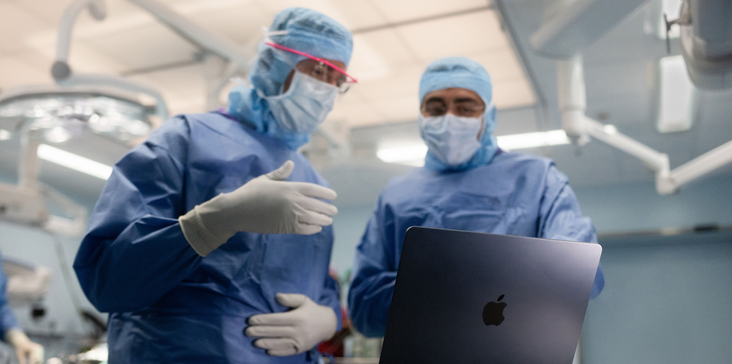 Dois cirurgiões na sala de operações olham para um MacBook; um faz gestos com a mão enquanto o outro observa