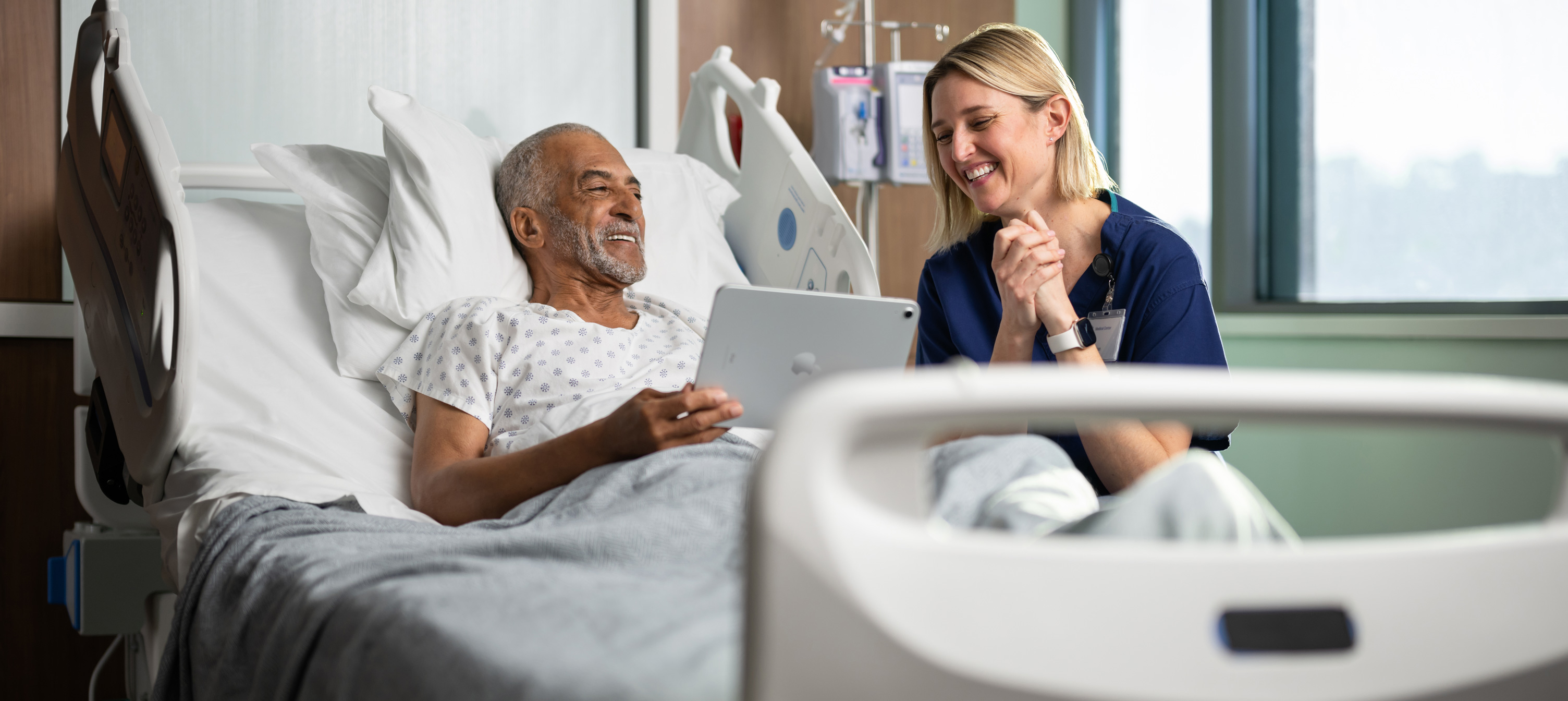 Een mannelijke patiënt zit in een ziekenhuisbed. Hij lacht en kijkt samen met een verpleegster naar een iPad-scherm.