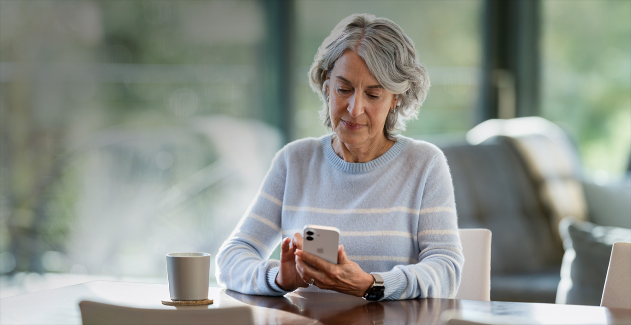 Een oudere vrouw zit aan een tafel en kijkt naar haar iPhone.