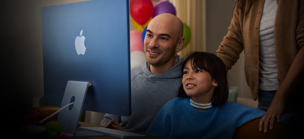 Birlikte oturan iki kişi bir iMac kullanıyor, üçüncü bir kişi arkalarında durarak onları izliyor.