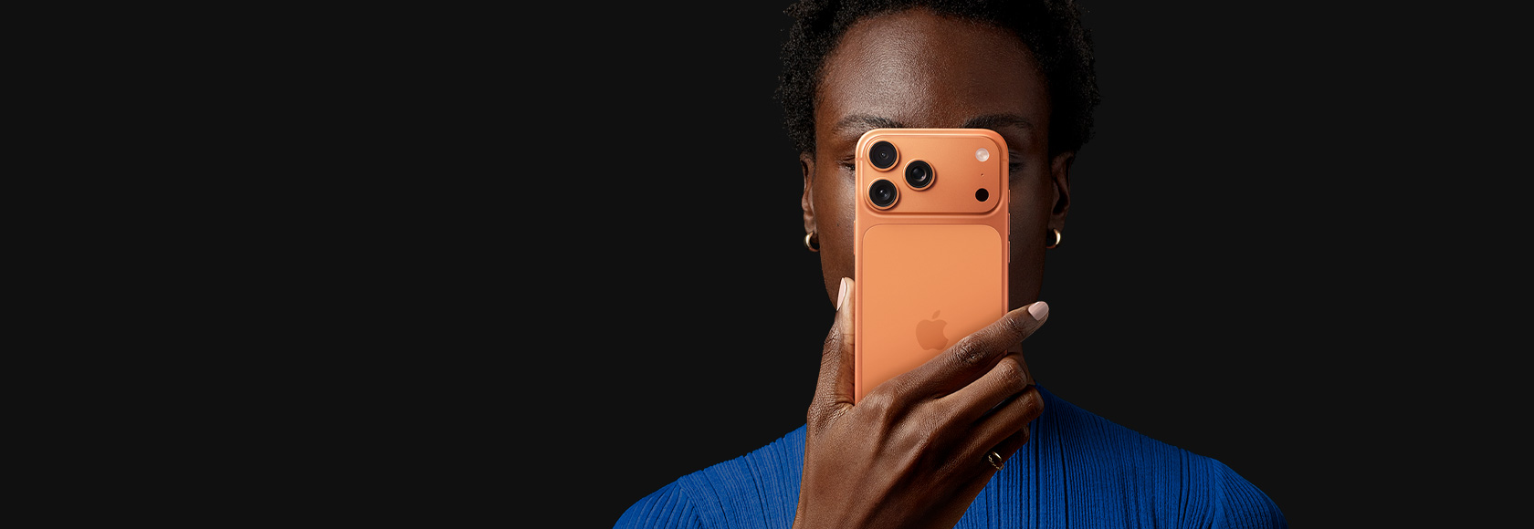 A person holds iPhone 17 Pro in front of their face, back exterior facing outward, Cosmic Orange color